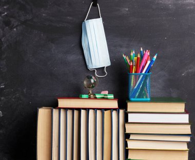 Kara tebeşir tahtasında asılı bir yığın kitap, kırtasiye malzemesi ve tek kullanımlık tıbbi maske, okula dönüş.