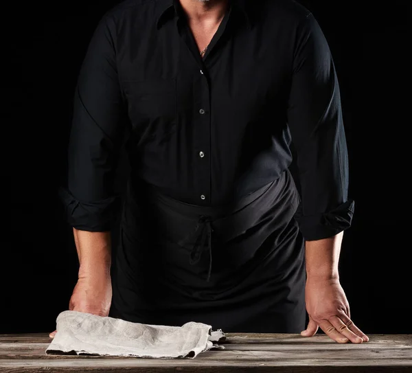 man chef in a black shirt and apron stands near a wooden table, rested his hands, black background