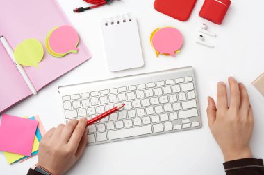 İki kadın eli ve bir beyaz klavye, açık bir defter, kulaklık, kırmızı akıllı telefon ve renkli çıkartmalar, beyaz masa, üst görünümü olan serbest çalışan işyeri.