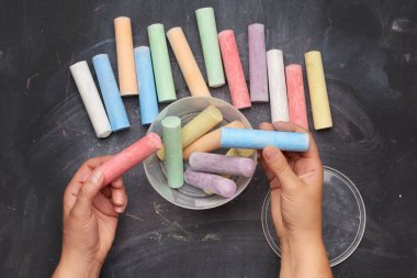 Siyah zemin üzerinde plastik bir kovada çok renkli tebeşirler, kadın elleri nesneleri bir kabın içine katlar, üst görünüm