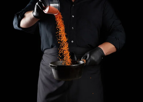 chef in black clothes and latex gloves pours raw lentils into a round cast iron pan with a handle, black background