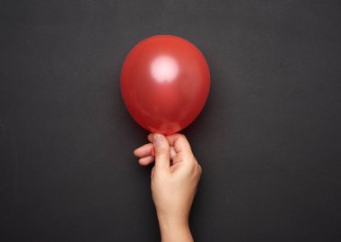 Kadın eli, siyah arka planda şişmiş bir kırmızı hava balonunu tutuyor. Yakın plan.