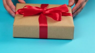 Kadın elleri kahverengi kraft kağıdına sarılı bir kutuyu tutuyor ve kırmızı ipek bir kurdele ile bağlanıyor, mavi arka plan.