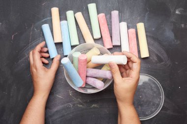 Siyah zemin üzerinde plastik bir kovada çok renkli tebeşirler, kadın elleri nesneleri bir kabın içine katlar, üst görünüm