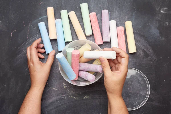 Siyah zemin üzerinde plastik bir kovada çok renkli tebeşirler, kadın elleri nesneleri bir kabın içine katlar, üst görünüm