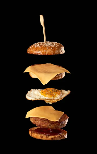 Uçan çizburger malzemeleri: et pirzolası, susamlı çörek, peynir, kızarmış yumurta ve ketçap, siyah arkaplan