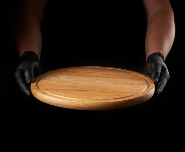 Siyah lateks eldivenli erkek elleri boş ahşap pizza tahtası ve siyah arka plan tutuyor.