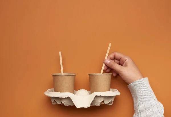 brown paper cups in a paper tray and a hand with a wooden stick on a brown background, no plastic, zero waste