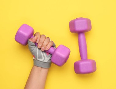 Pembe spor eldivenli kadın eli sarı arka planda mor bir kilogram dambıl tutuyor.
