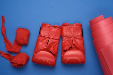 Bir çift kırmızı deri boks eldiveni ve tekstil kırmızısı elastik bandaj. Eller mavi arka planda, spor setinde, düz yerde yatar.
