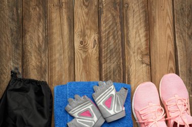 Bir çift pembe spor ayakkabı, mavi havlu ve spor eldivenleri gri ahşap arka planda, üst manzara