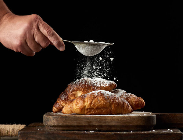 man's hand holds a metal strainer with powdered sugar and sprinkles baked crispy croissants on a brown wooden board, black background