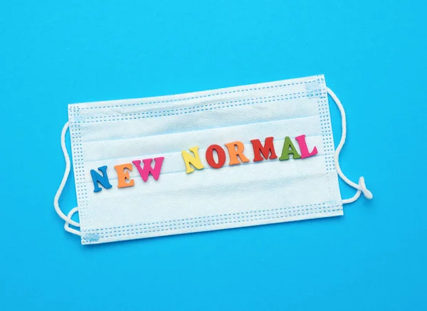 disposable medical face mask and lettering new normal on blue background, concept of wearing protective equipment during a pandemic, flat lay