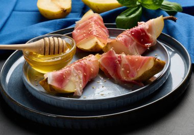 Bir armut aperatifi Prosciutto, susam tohumları serpiştirilmiş ve altın bal ile servis edilmiş. Tabak mavi bir tabakta taze fesleğen yapraklarının yanında servis edilir..