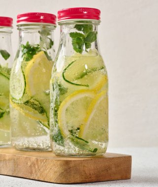 Kırmızı kapaklı cam şişeler, içinde salatalık, limon dilimleri ve nane yaprakları bulunan detoks suyla dolu beyaz masa.