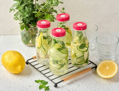 Kırmızı kapaklı cam şişeler, içinde salatalık, limon dilimleri ve nane yaprakları bulunan detoks suyla dolu beyaz masa.