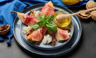 Prosciuttoya sarılmış armut parçaları ve susam tohumları serpiştirilmiş, küçük bir bal kasesi ve mavi seramik tabakta ahşap bir bal kaşığı ile servis edilmiş..