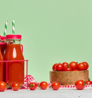 Kırmızı metal bir stantta domates suyuyla doldurulmuş kırmızı başlıklı cam şişeler. Taze kiraz domatesleri ve yakınlarda bir yeşillik filizleri var.
