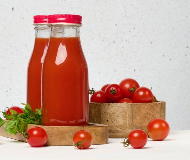 Kırmızı başlıklı domates suyu ile dolu cam şişeler. Taze kırmızı domatesler ahşap bir standın üzerinde ve önlerinde beyaz bir zemin var.