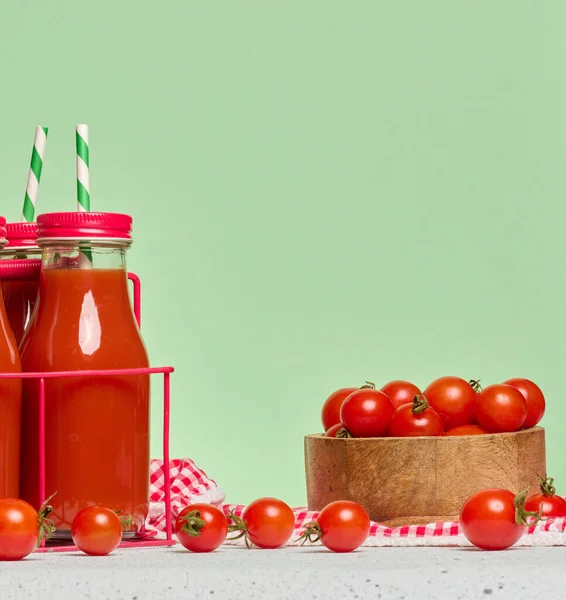 Kırmızı metal bir stantta domates suyuyla doldurulmuş kırmızı başlıklı cam şişeler. Taze kiraz domatesleri ve yakınlarda bir yeşillik filizleri var.