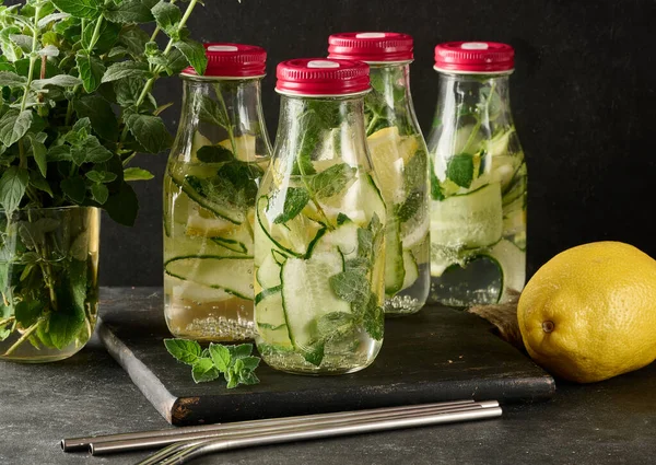 İçinde salatalık, limon dilimleri ve nane yaprakları bulunan kırmızı kapaklı cam şişeler. Şişeler ahşap bir tahtanın üzerinde, arka planda koca bir limon var.