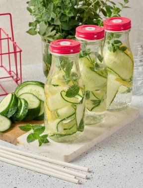 Kırmızı kapaklı cam şişeler, içinde salatalık, limon dilimleri ve nane yaprakları bulunan detoks suyla dolu beyaz masa.