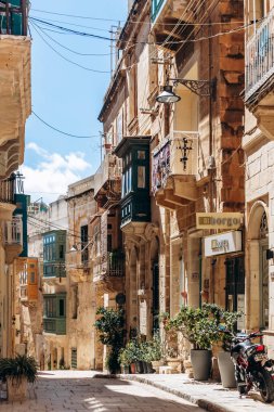Senglea, Malta - 11 Nisan 2025: Senglea 'da sakin tarihi bir sokakta güneşli bir bahar günü geleneksel Malta kireçtaşı cepheleri, renkli ahşap balkonlar, saksı bitkileri, dükkan işaretleri