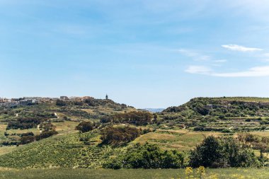 Yeşil, yuvarlanan manzaranın panoramik manzarası ve Gozo adasının tipik tarlaları, köy binaları ve açık bir yay gökyüzünün altındaki uzak tepede bir kilise kubbesi..