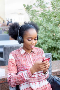 Kulaklıklar açık hava kafede otururken, smartphone içinde yazarak ve gülümseyen boyun ile etnik elbiseli siyah afro kız.