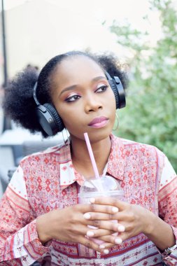 Müzik ve süt kokteyl içme siyah afro kız açık hava kafede oturan kulaklıklar ile etnik elbiseli