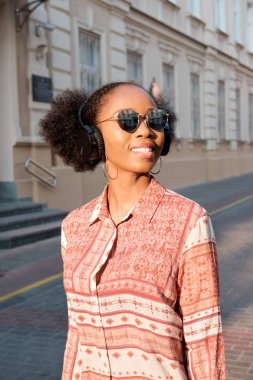 Siyah afroamerican kız akşam kasaba ve kulaklık müzik dinleme bir yürüyüşü vardır. Kız güneş gözlüğü yaz gülümsüyor ve sunset Bulvarında arıyor.