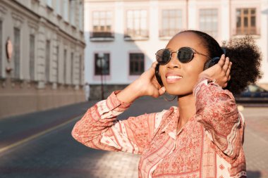 Siyah afroamerican kız akşam kasaba ve kulaklık müzik dinleme bir yürüyüşü vardır. Kız güneş gözlüğü yaz gülümsüyor ve sunset Bulvarında arıyor.