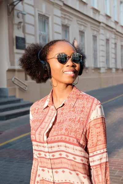 Siyah afroamerican kız akşam kasaba ve kulaklık müzik dinleme bir yürüyüşü vardır. Kız güneş gözlüğü yaz gülümsüyor ve sunset Bulvarında arıyor.