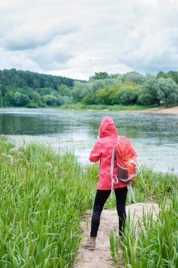 Young woman, weared in pink raincoat, with orange travel backpack on a shoulder, stands on a bank of river and observes faraway landscape. Concept travel. clipart