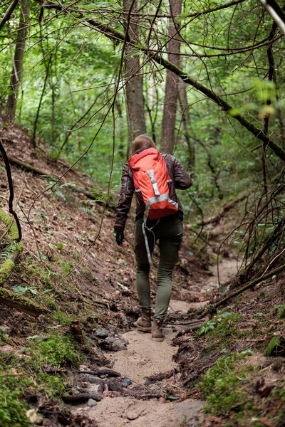 Young redhead long hair woman travels with red travel backpack in summer cloudy forest. Concept travel.