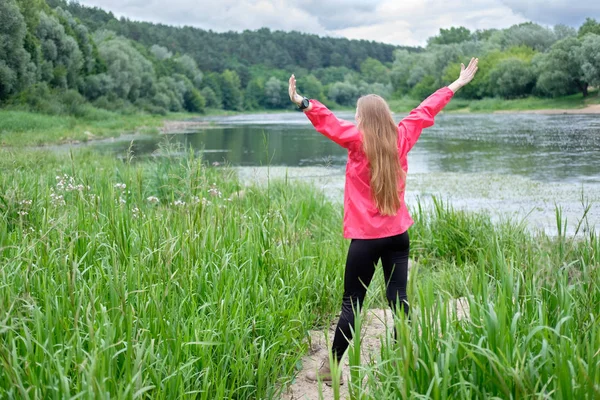 Young woman, weared in pink raincoat, stands on a bank of river and observes faraway landscape, rising hands up. Concept travel.