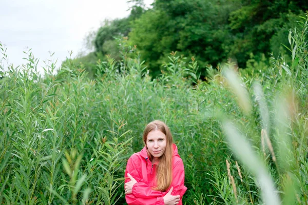 Kızıl saçlı Genç kadın, pembe yağmurluk renksizdir donuyor ve kendini yeşil çalıların ayakta sarılarak. Yaz bulutlu yağmurlu hava.