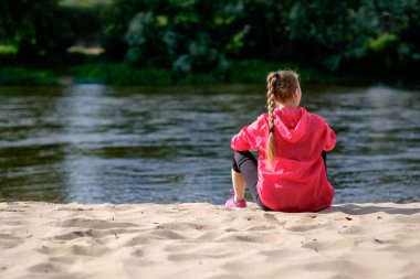 Kızıl saçlı beyaz sıska girk ile örgü, pembe yağmurluk, kumlu bir nehir kıyısında oturan renksizdir, arkadan görüntüleyin. Sıcak güneşli yaz hava.