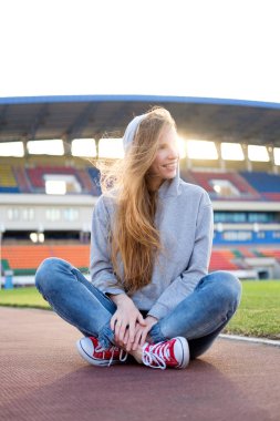 Kızıl saçlı beyaz genç kadın oturuyordu ve çim futbol sahası ve akşam günbatımında hafif gülümseyerek ralaxing. Arka plan üzerinde stadyum standları.