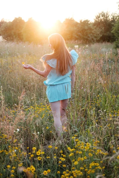 Young redhead woman in blue dress walking in sunset field in summer.