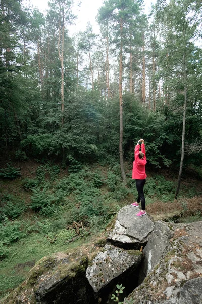 Young woman in pink raincoat enjoys fresh forest air, standing on a top of a rock.