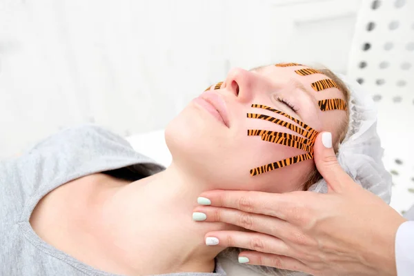 Cosmetologist making taping face procedure using tiger colored tapes in beauty parlor. Patient lying on the procedure table and smiling. Concept beauty.