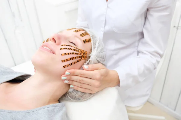 Cosmetologist making taping face procedure using tiger colored tapes in beauty parlor. Patient lying on the procedure table and smiling. Concept beauty.