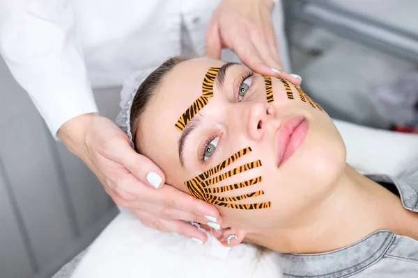 Cosmetologist making taping face procedure using tiger colored tapes in beauty parlor. Patient lying on the procedure table. Concept beauty.