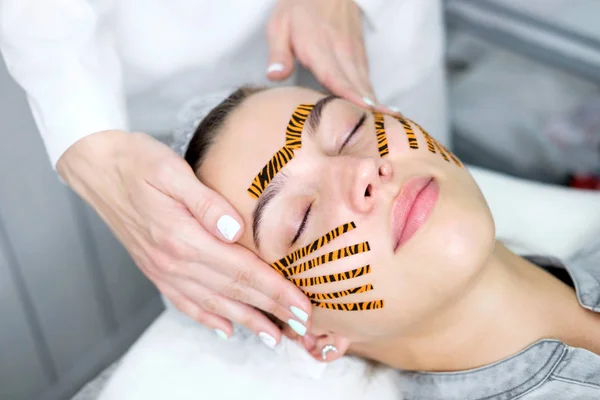 Cosmetologist making taping face procedure using tiger colored tapes in beauty parlor. Patient lying on the procedure table. Concept beauty.