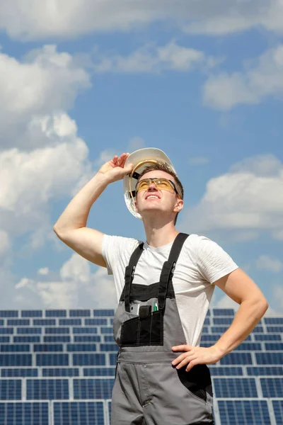 Solar panels engineer in white cask, protective yellow glasses and grey outfit stadnding near solar panels field and watching at the sun. Hot sunny summer weather. Blue sky background. Concept of renewable and clean energy, electricity, technology.