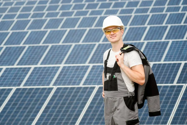 Solar panels engineer in white cask, protective yellow glasses and grey outfit stadnding near solar panels field and smiling. Hot sunny summer weather. Copy space. Concept of renewable and clean energy, electricity, technology.