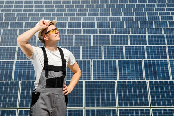 Solar panels engineer in white cask, protective yellow glasses and grey overalls stadnding near solar panels field. Hot sunny summer weather. Copy space. Concept of renewable and clean energy, electricity, technology.