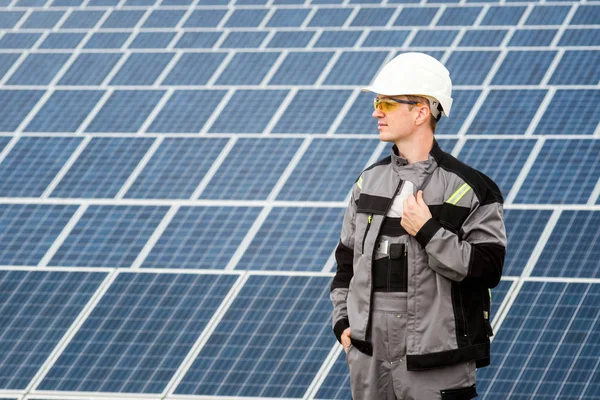 Solar panels engineer in white cask and grey outfit stadnding near solar panels field. Hot sunny summer weather. Copy space. Concept of renewable and clean energy, technology.