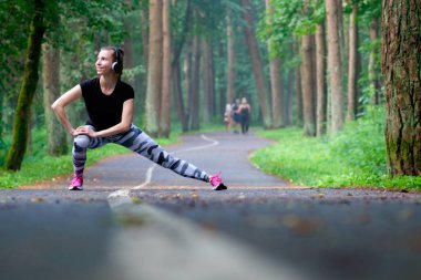 Güzel çekici genç kadın parkta, germe dinle müzik ve gülümseme. İnsanların arka plan üzerine bir yürüyüş var. Yaz saati. Sağlık kavramı. Fitness kavramı.